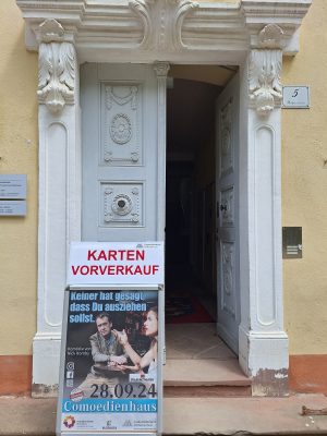 Eingangstür zur Geschäftsstelle des Comoedienhauses in Wilhelmsbad. Davor steht ein Plakataufsteller mit Ankündigung des Schauspiels Keiner hat gesagt, dass du ausziehen sollst